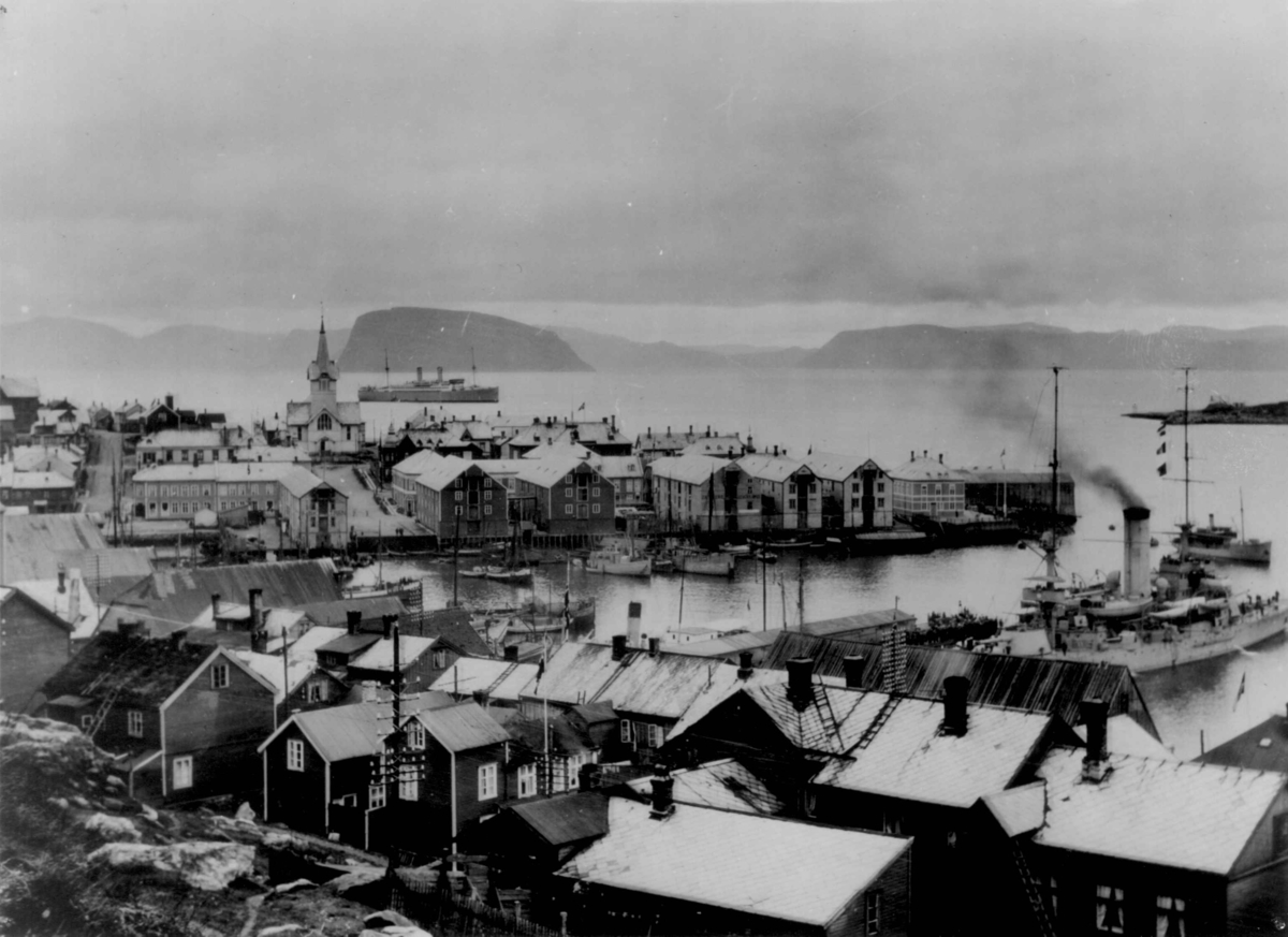Utsikt over Hammerfest med byen og havna. Trehus, boliger og sjøboder med en kirke i bakgrunnen. Et skip ligger ved kai, ute på fjorden sees flere båter.