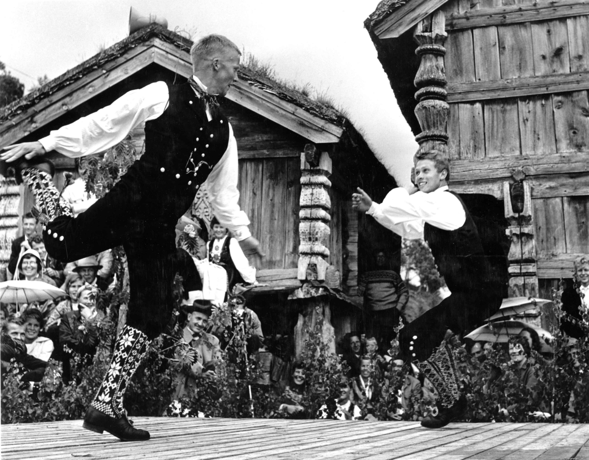 To hallingdansere i dans på scene i Friluftsmuseet på Folkemuseet. Tilskuere foran scena og tømmerbygninger i bakgrunnen. Setesdalstunet.