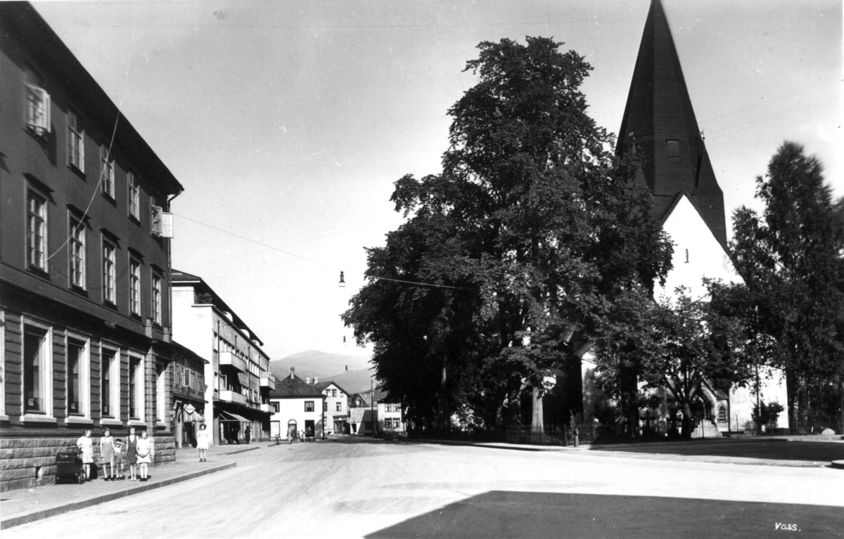 Voss kirke 1939.  Såvidt synlig bak trær. Gate til venstre. Småjenter foran et hus.