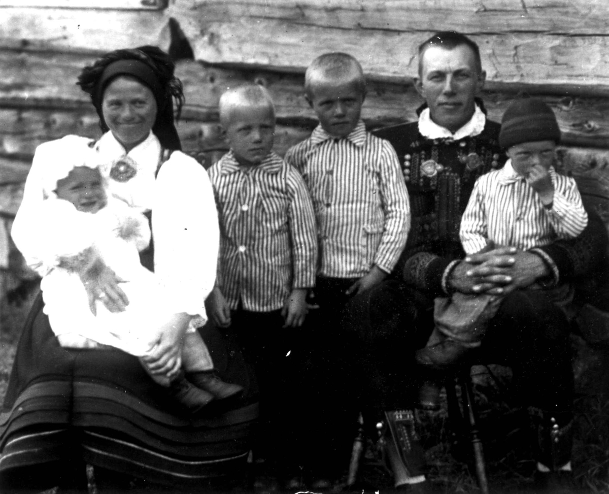 Kvinne-, manns- og barnedrakt, gruppeportrett, Valle, Setesdal, Aust-Agder, antatt 1924. Familiegruppe sittende foran tømmervegg. Fra "De Schreinerske samlinger" (skal oppgis).
