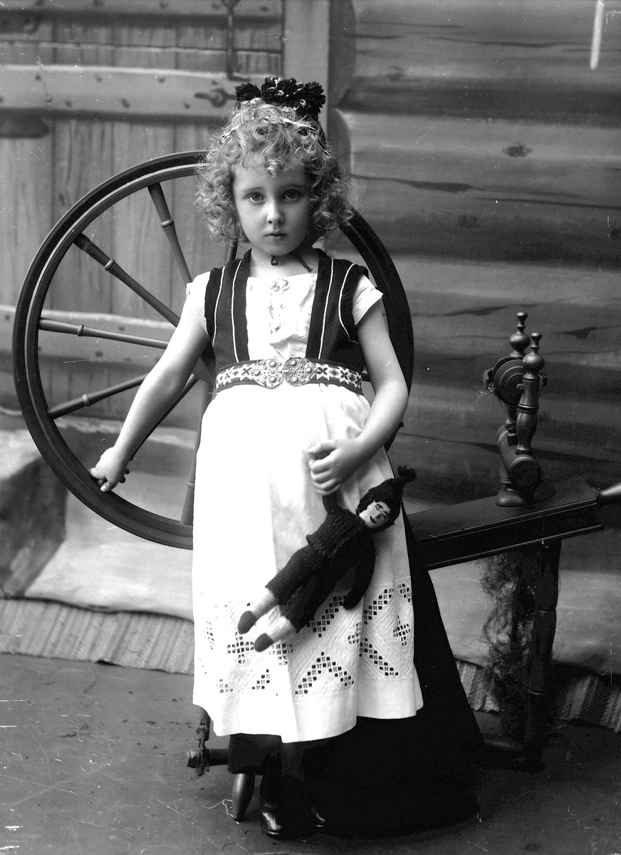 Studioportrett av jente i nasjonaldrakt som lener seg mot hjulet på en rokk. Jenta holder en dukke i hånden og ser på betrakteren. Bakgrunnen er malt og viser trevegger.