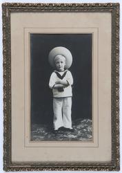 Portrett av Kronprins Olav i hvit matrosdress og stor hatt.