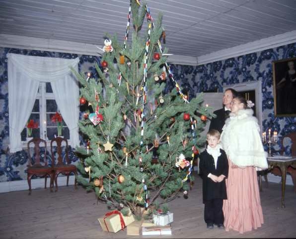 Det feires jul i Leikanger prestegård, bygning nummer 191 på Norsk Folkemuseum.Museumsansatte  samt et barn, alle i kostymer beundrer det nytente juletreet.