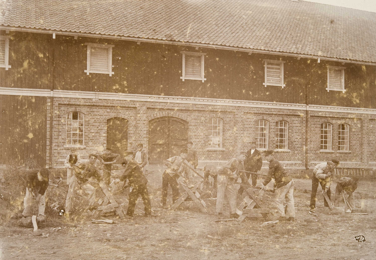 Elever utenfor låvebygningen på Bastøy skolehjem, Bastøya, ca. 1903. Elevene sager ved.