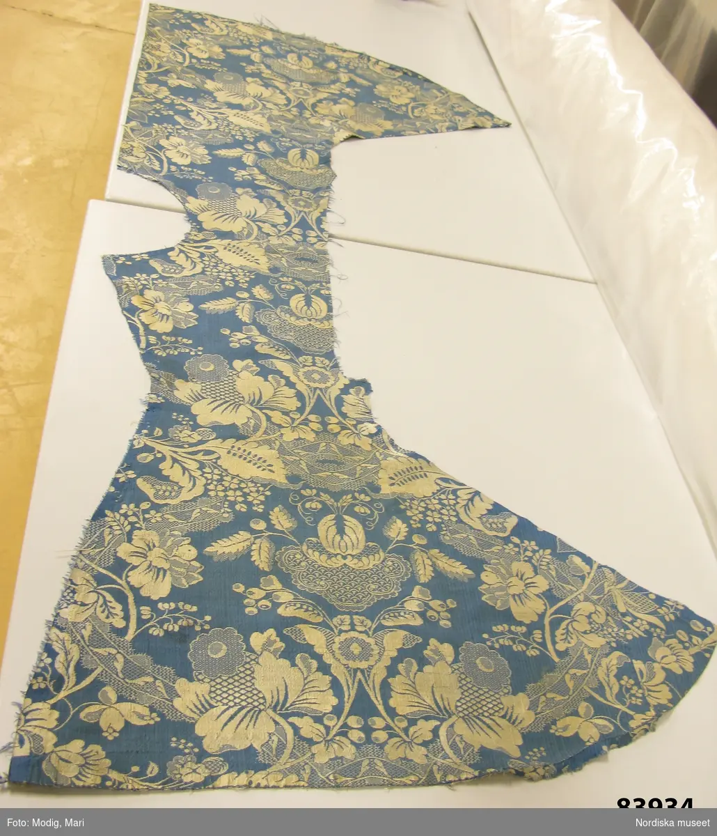 Kvinnokofta av mönstervävt siden, blå botten med blom-och bandmönster i vitt. Skuren i 2 delar. Nu uppsprättad och med delvis bortskurna bitar.
/Berit Eldvik 2013-01-09