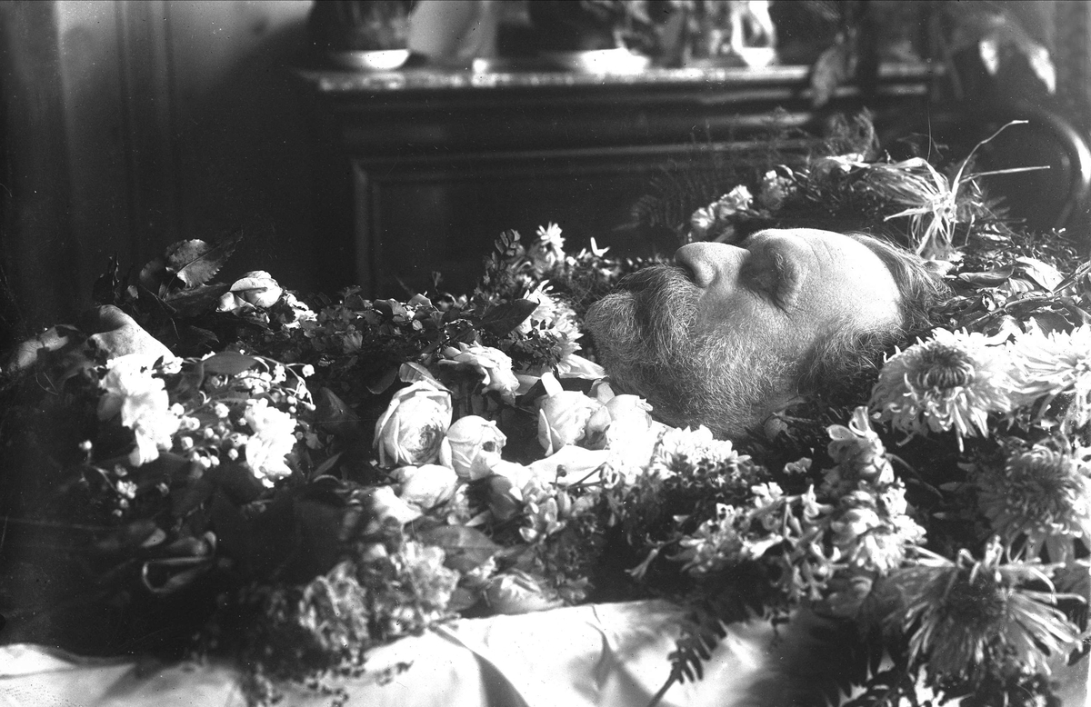 Post mortem-fotografi. Død mann i kiste pyntet med blomster.