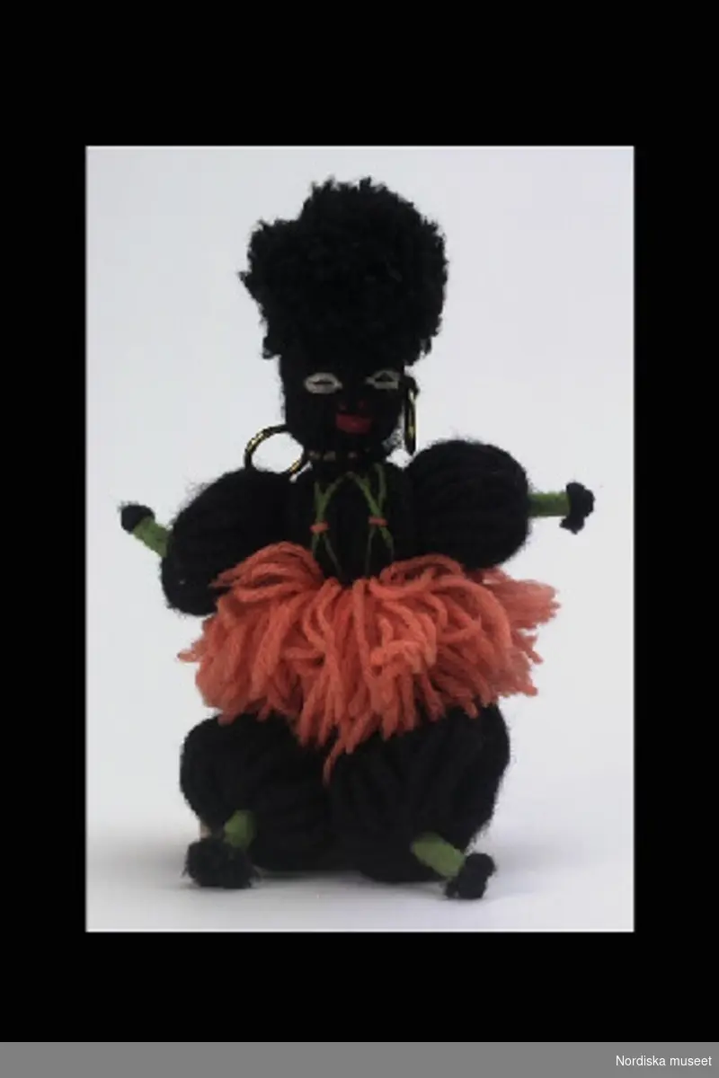 Inventering  Sesam 1996-1999:
Docka av svart ullgarn med sydda ögon och mun, kjol av orange garn, gröna händer och fötter, ringar av mässing i öronen.
/Leif Wallin 1996