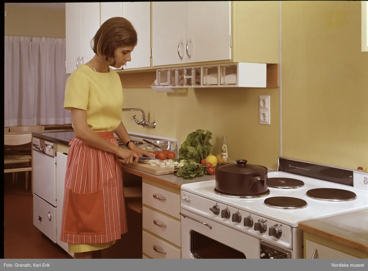 Köksinteriör med kvinna i randigt förkläde som arbetar vid köksbänken. Hon skär grönsaker på en skärbräda medan en gryta står på spisen.