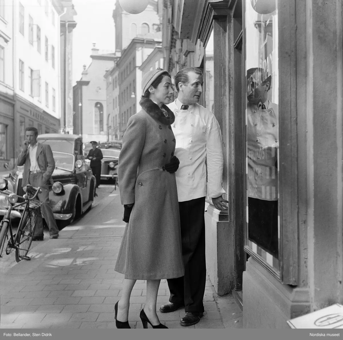 Modebild för Sahlins konfektionsfabrik. Modell i kappa med pälskrage, högklackat och hatt står tillsammmans med en kypare och tittar in i fönstret till restaurang Cattelin, Gamla stan, Stockholm.