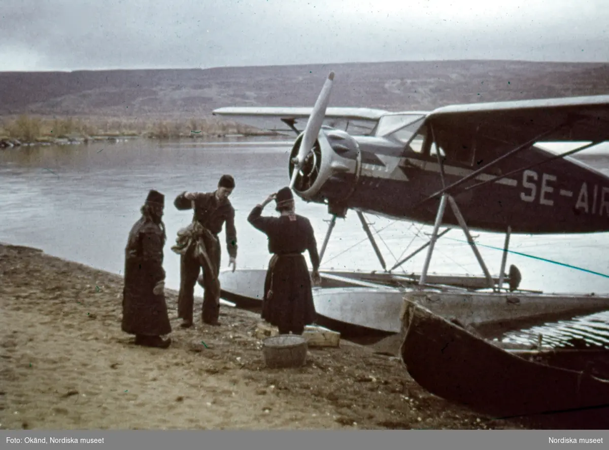 Påskrift på monteringen: "Fiskflygare vid Sitojaure." Två samer och pilot vid sjöflygplan som landat vid stranden intill en uppdragen båt.