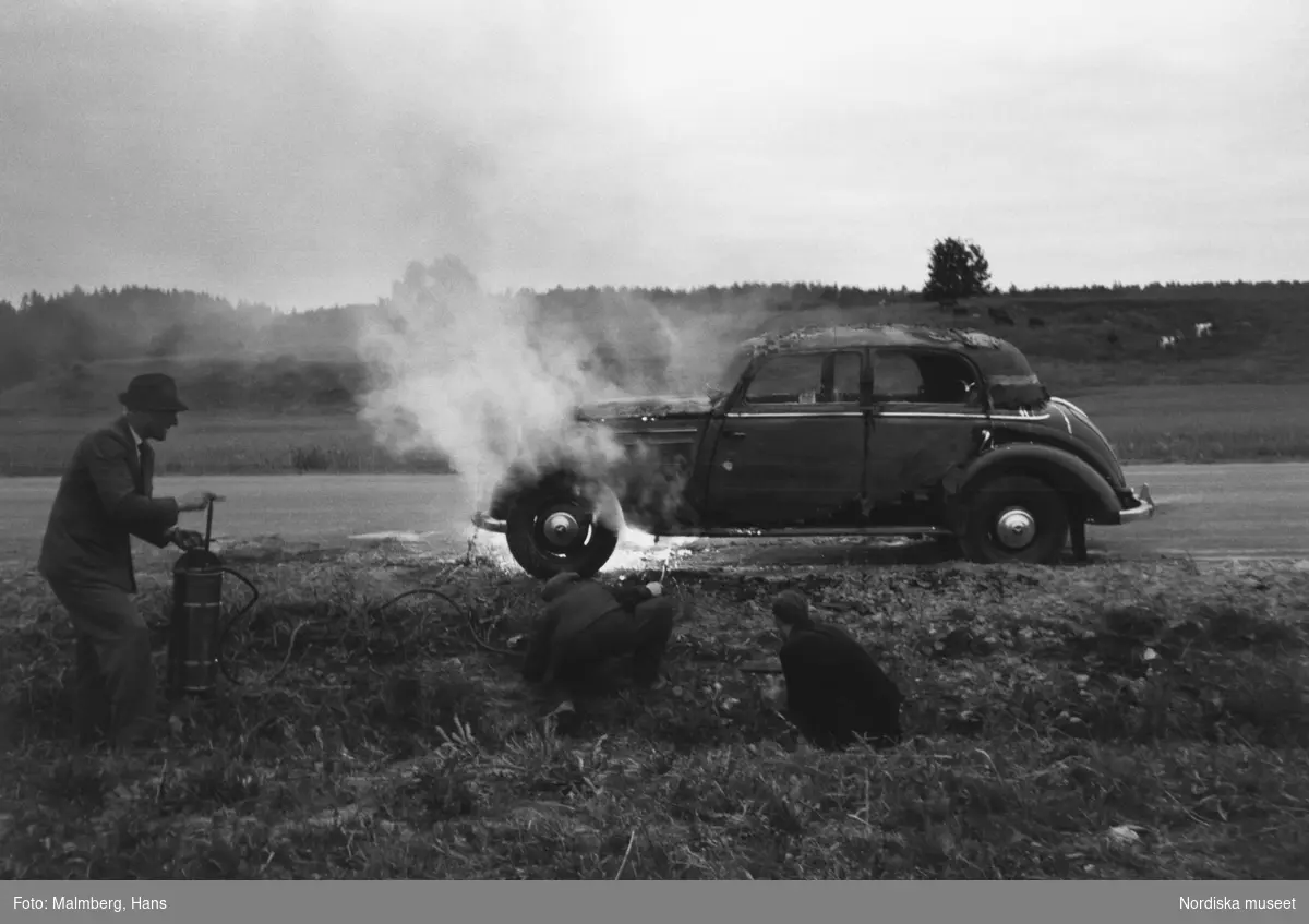 På Riks-Ettan 1954. 
Tre män försöker släcka en bilbrand vid vägkanten.
