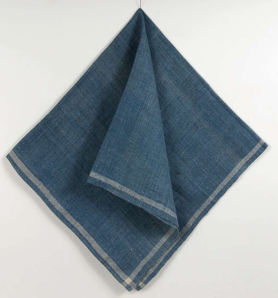 Halskläde av blå kypertvävd linne med vit rand runt kanten. I ett hörn märkt I O med blå korsstygn. Fållad på två sidor, på den ena ett blått band fastsytt på baksidan efter märkningen.