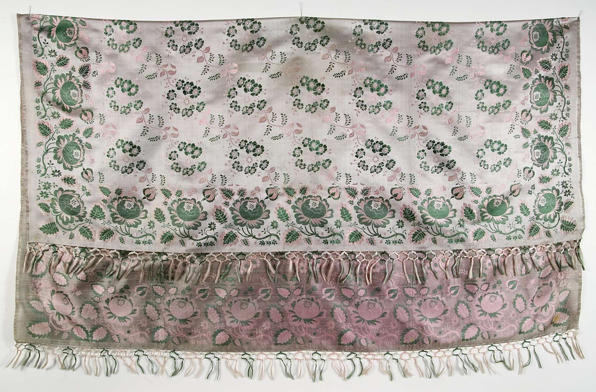 Kläde av ljusgrått siden med invävt blommönster i rosa, grönt. Iknuten frans på varpsidorna. Stockholms hallstämpel 1842.