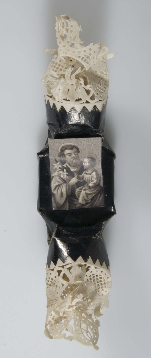 Begravningskaramell, svart glanspapper och vitt tårtpapper. Svartvit påklistrad bild: manligt helgon och Jesusbarnet.