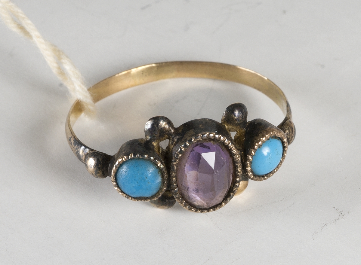 Fingerring. Troligen förgyllt silver. Smal ring med tre stenar, oval violett fasettslipad ametist, omgiven av två runda turkoser.
Inga stämplar.
