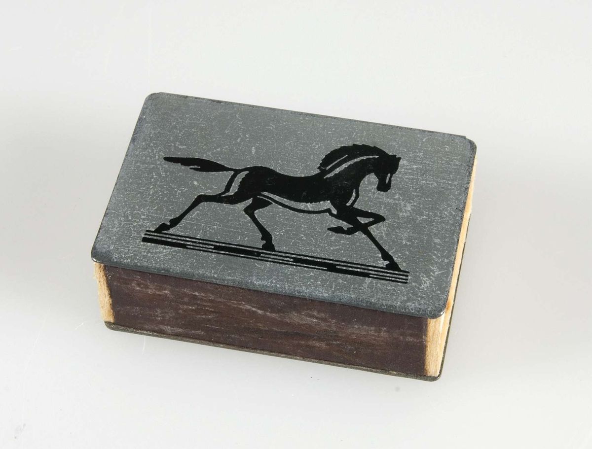 Fodral i vitmetall till tändsticksask. Text i svart: Hästens Skor. På andra sidan bild av häst. Innehåller liten tändsticksask med stickor.