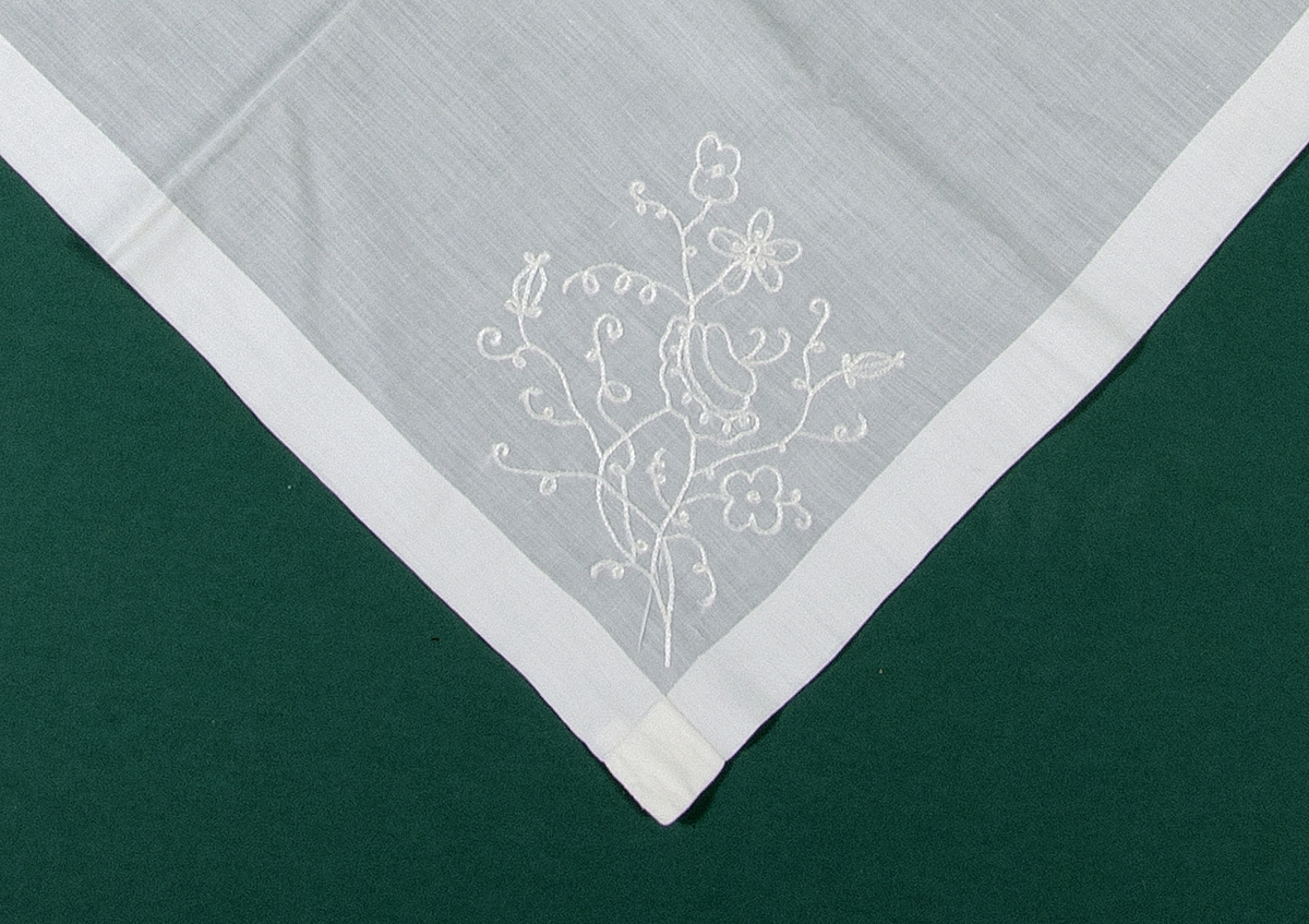 Schalett, handsydd av tunt vitt bomullstyg vävt i tuskaft och med broderade blommor i två av hörnen. Broderierna är gjorda med kedjestygn och vitt silkesgarn.