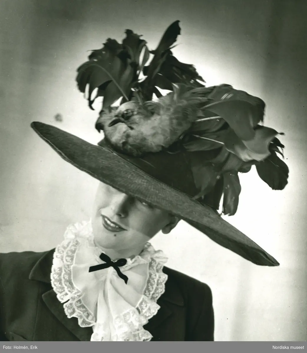 Porträtt av kvinna i bredbrättad hatt med fjädrar och fågeldekoration (NM.0213374), av Suzanne Pellin, samt vit spetskrage och mörk kavaj.