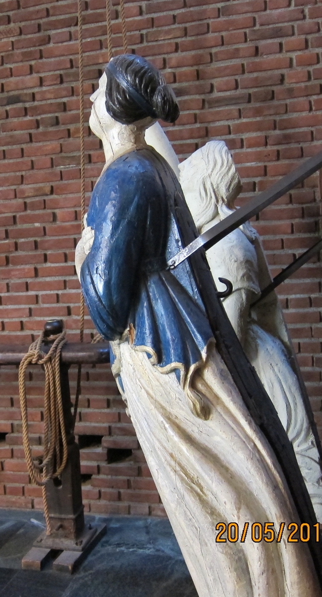 Kvinne med armene i kryss over brystet, blå overdel, hvit kjole med gullkant som pynt, sort hår med hårbånd.