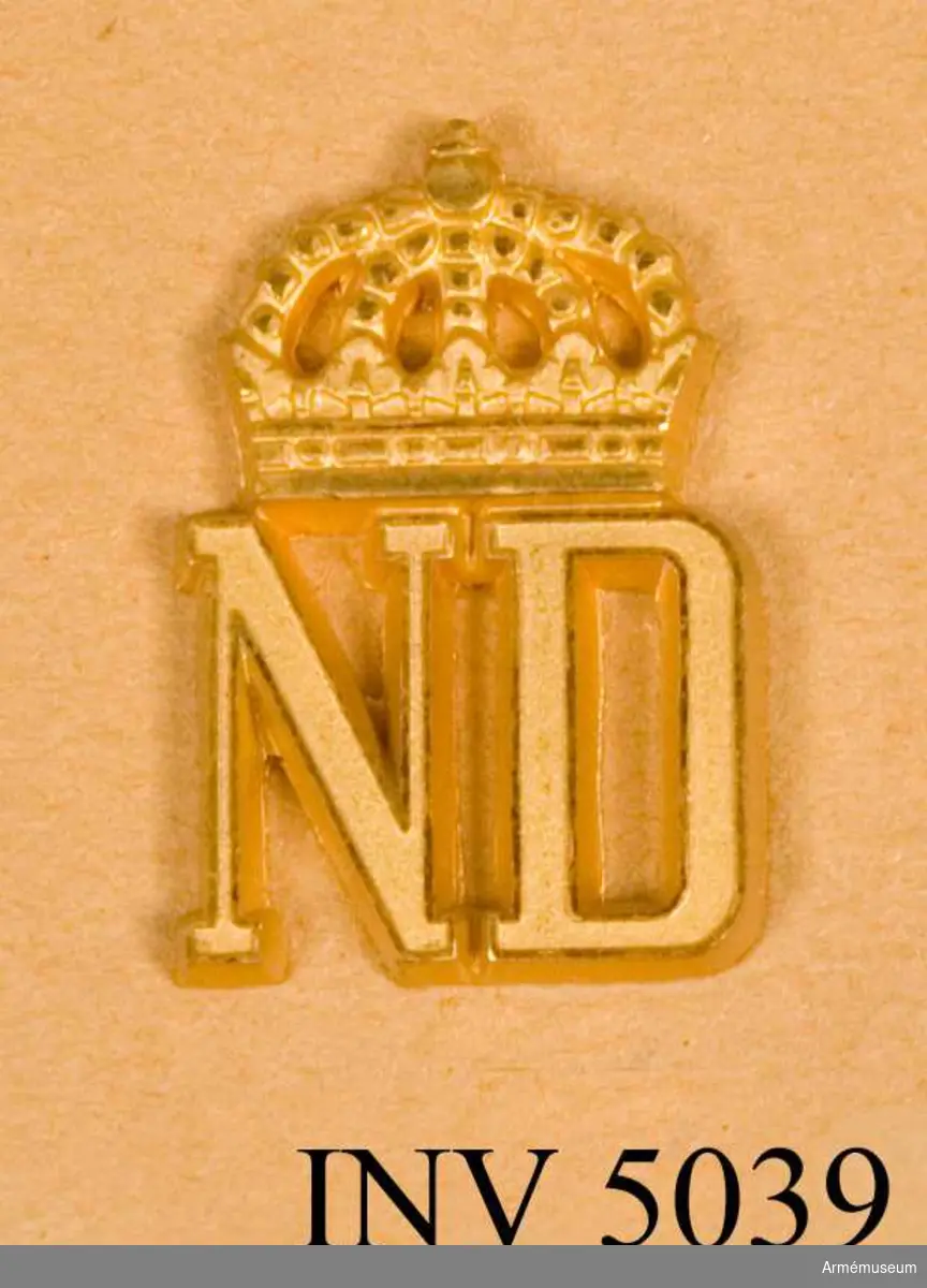 Förbandstecken m/1961, Norrlands dragoner.Tillverkat av metall och i metallfärg m/1960 guld.Till uniform m/ä hade regementet som förbandstecken bokstäverna NDR under kungl krona. Till övriga uniformer förbandssiffror. Före 1928 hade regementet nummer 8, men erhöll detta år nummer 4 i samband med att Smålands husarregemente indrogs. Den 11 juli 1957 fastställdes nytt förbandstecken. Det blev en återgång till det gamla tecknet men medtogs inte bokstaven R enär regementet skulle omorganiseras till bataljon och fick förbandet då också sitt nuvarande namn. Ädelsmeden Sigurd Persson har vid sin formgivning av tecknet i huvudsak behållit 1957 års modell, se 4694, men något förändrat bl a kronan. Anläggande och anbringande se 4698.Vidhängande etikett anger dag för fastställande. Stockholms Slott den 25/4 1962  Sven Andersson H. Henning .