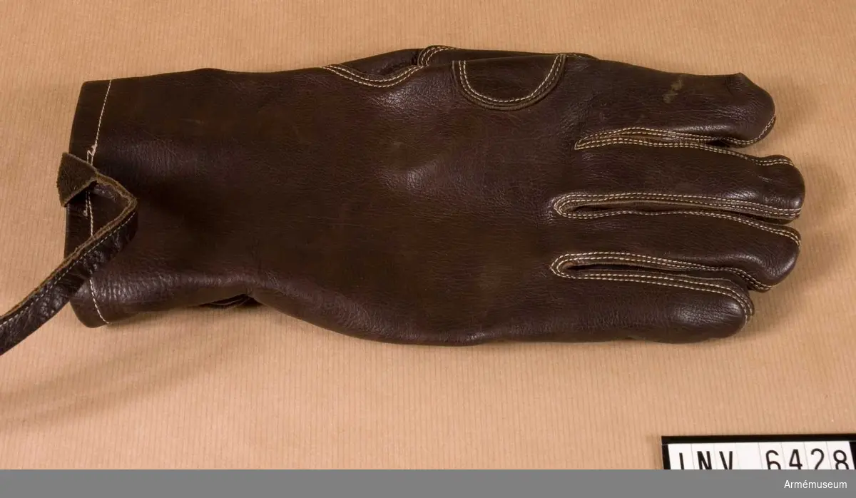 Handske m/1929, ytter- f kavalleri. Stl -. Av mörkbrunt läder m rem f fastsättning vid handleden och rem f åtdragning vid handleden. Extra förstärkning i handflatan. Sömmen är ljus.