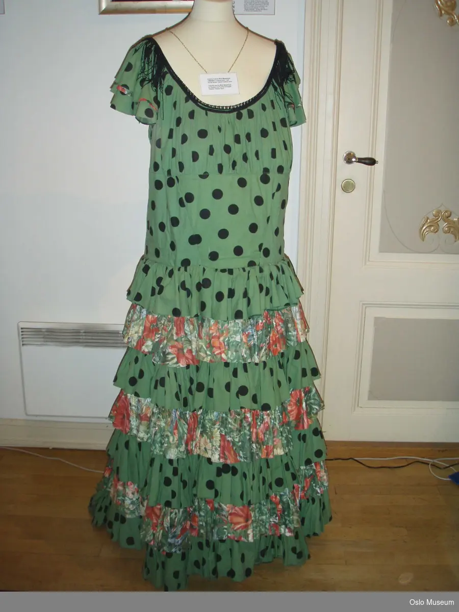 A= Lang kjole i bomull, grønn med sorte prikker og rysjer og kapper.
B= Underskjørt i lerretsstoff.