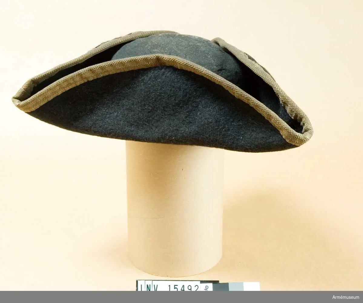 Trekantigt uppviken hatt av svart filt med vitt ylleband i kanten.