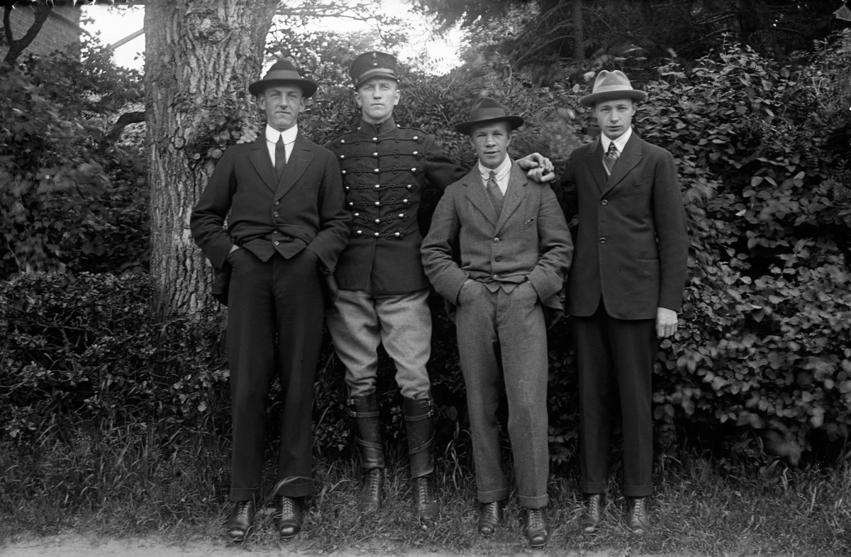 Fyra män, en klädd i uniform och Gustav Andersson längst till höger, står uppställda på rad med träd och buskar som bakgrund.