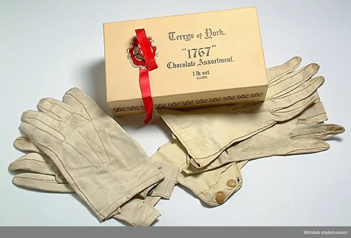 Vita glacéhandskar, tre par och en udda handske förvarade i en chokladask.