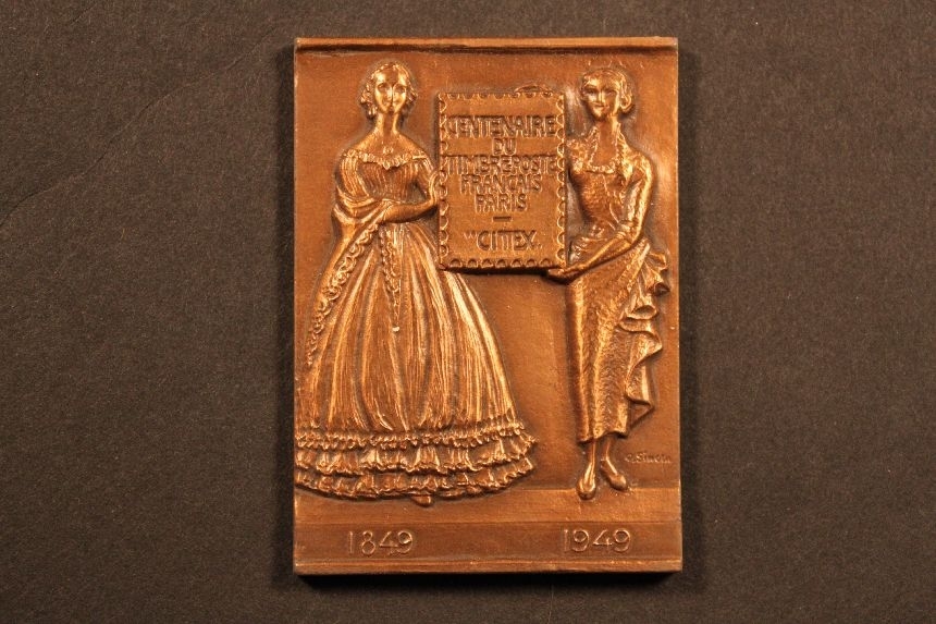 Plakett i brons, rektangulär. Motivet visar två kvinnor
idräkter från 1849 respektive 1949, vilket också anges
underkvinnorna. De båda håller i en skylt, utformad som ett frimärke.
Påskylten står text enligt MRK. Baksidan är blank.