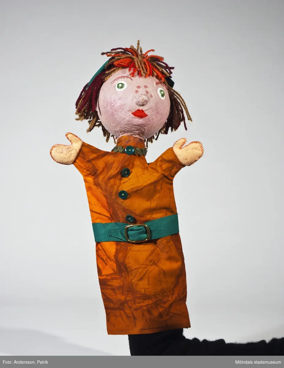 Greta ur sagan om Hans och Greta (se 04905).Handdockan har ett huvud av gips, brunt och rött hår av garn med en grön rosett, en "klänning" av mönstrat orange tyg med gröna knappar och ett grönt bälte med spänne av mässing.