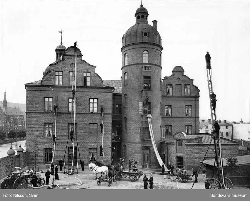 Gamla brandstationen. Brandredskapen demonstreras under ledning av brandchef Gustaf Hellgren som står längst ned till höger. Stora ångsprutan till vänster drogs av hästar.
