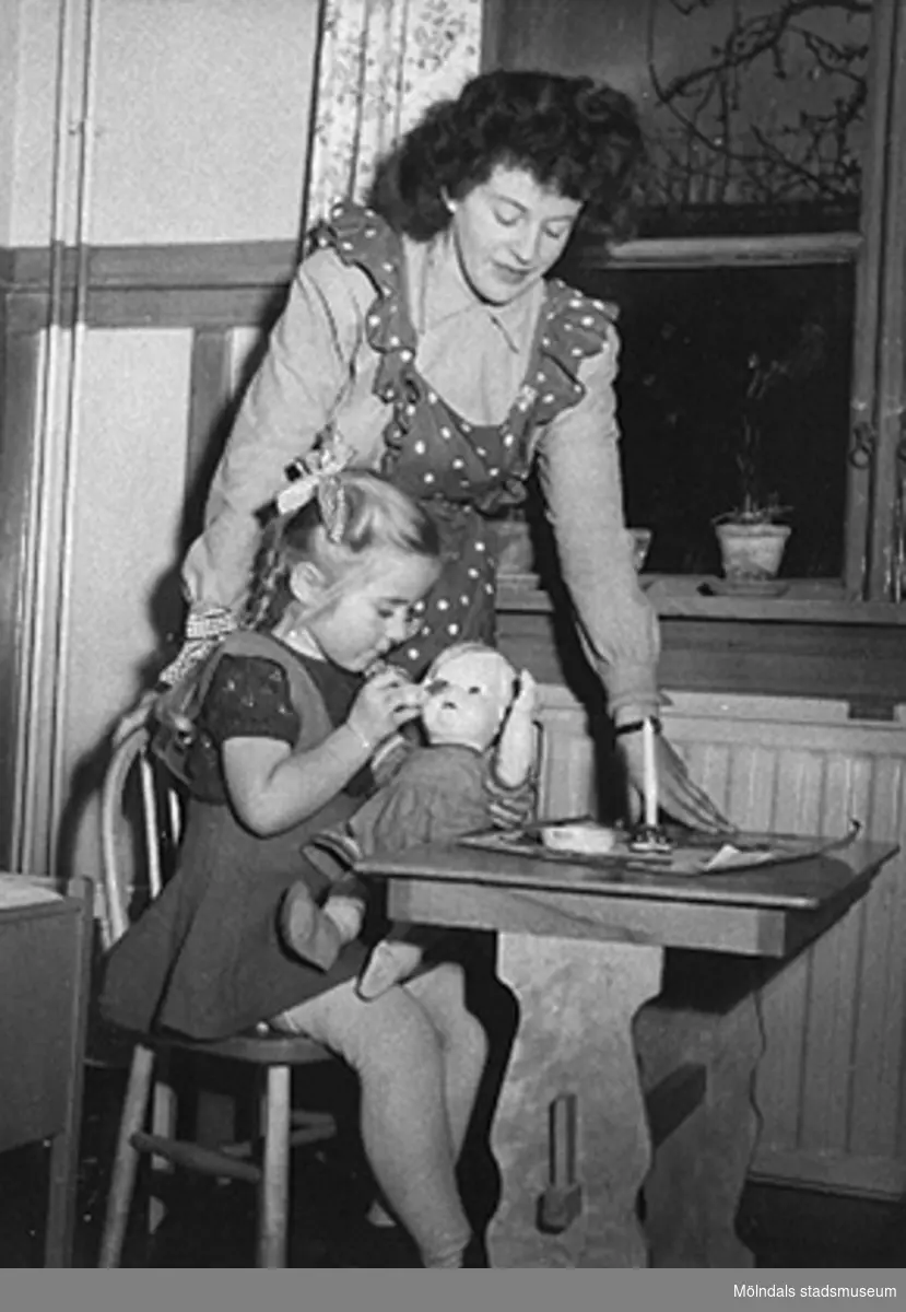 Bitr. föreståndare Margit Emilsson (gift Wannerberg -52) tittar på när en flicka pysslar om en docka, Krokslätts daghem år 1949.

Margits rödprickiga förkläde är av samma tyg som gardinerna bakom henne. Tyget är från Mölnlycke fabriker.