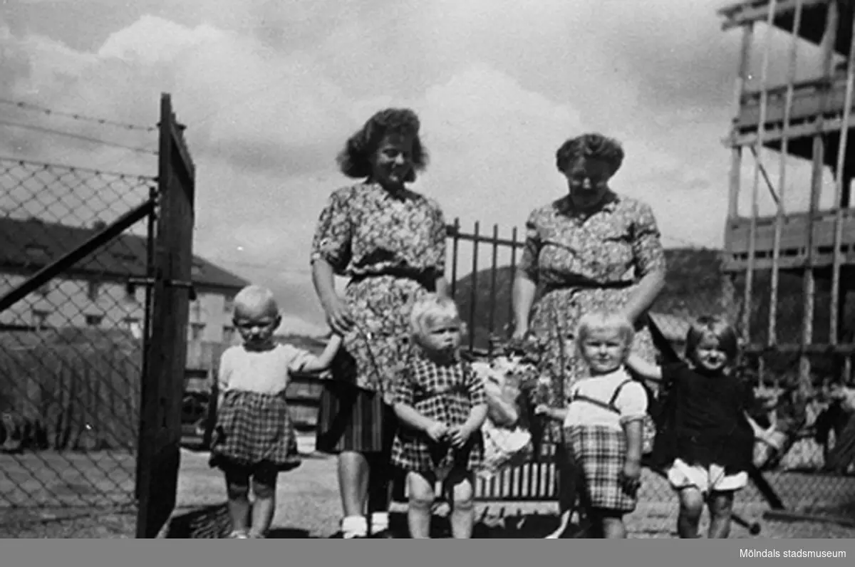 Praktikant Margit Emilsson (gift Wannerberg -52) och en lärare står utomhus tillsammans med fyra barn vid Krokslätts daghem. 
Under Margits praktik som biträde 1945-10-01 - 1946-08-01.
