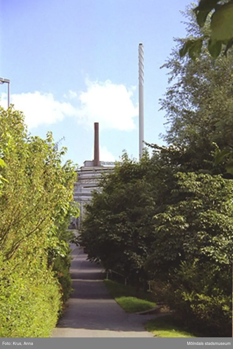 Asfalterad gångväg, grönska, takåsar och två skorstenar fotograferade nerifrån gångbanan vid Kvarnbygatan i Mölndals Kvarnby. Den vänstra röda skorsten sitter på "Strumpans" tak (Kvarnbygatan 10-14) och den högra vita skorstenen sitter på den s.k O-fabriken. O-fabriken tillhörde tidigare SOAB och beteckningen står för Oljefabrik. Byggnaden har inrymt många olika funktioner som varierat allt eftersom fabrikens tillverkning förändrats, bl a panncentral (därav skorstenen).