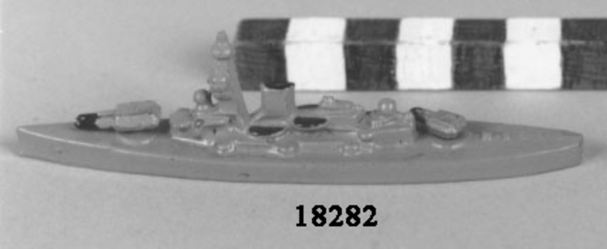 Fartygsmodell i form av pansarskeppet Gustav V gjuten av metall i ett stycke, målad i grått. Plan botten. Artilleripjäser i för och akter, dubbla vridbara. Mast och livbåtar med svart kapell, fyra stycken.