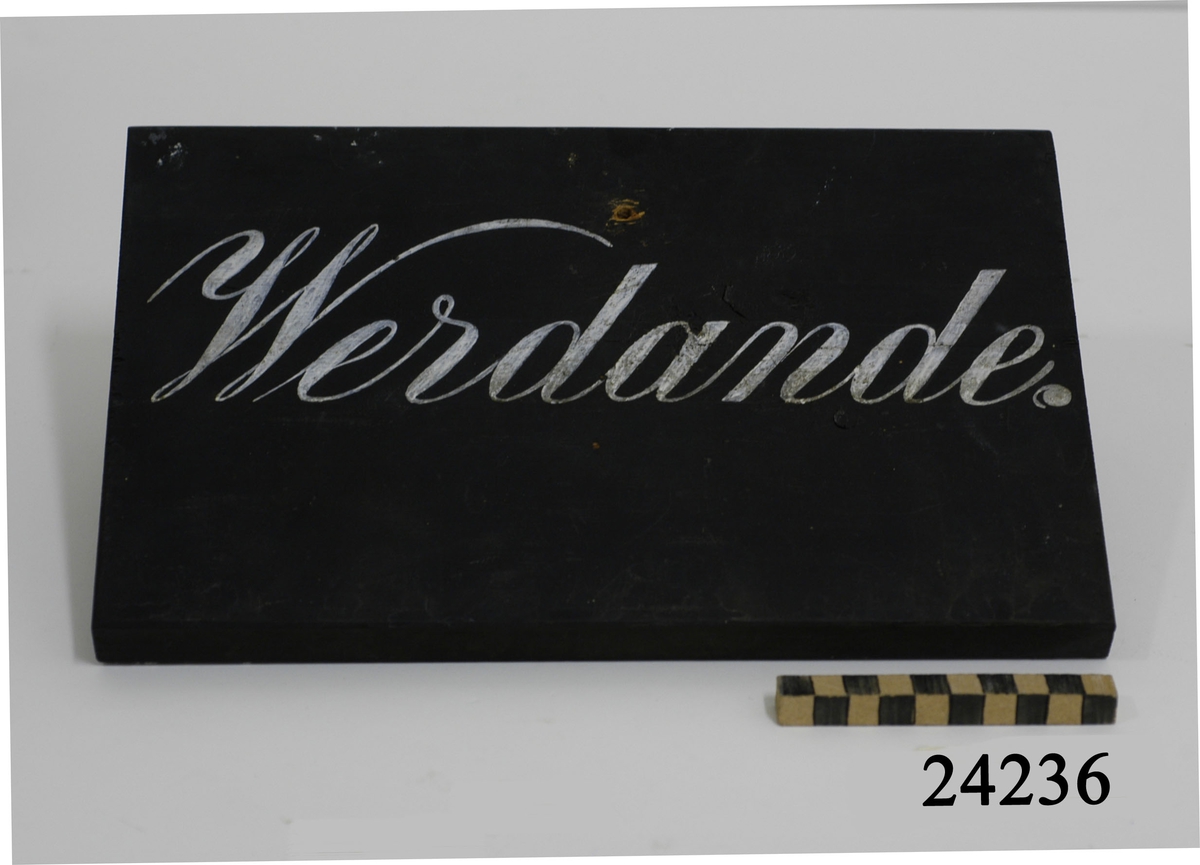 Uppbördsskylt av trä, svartmålad med vit handmålad text: "Werdande". I skyltens övre kant ett borrat hål för upphängning.