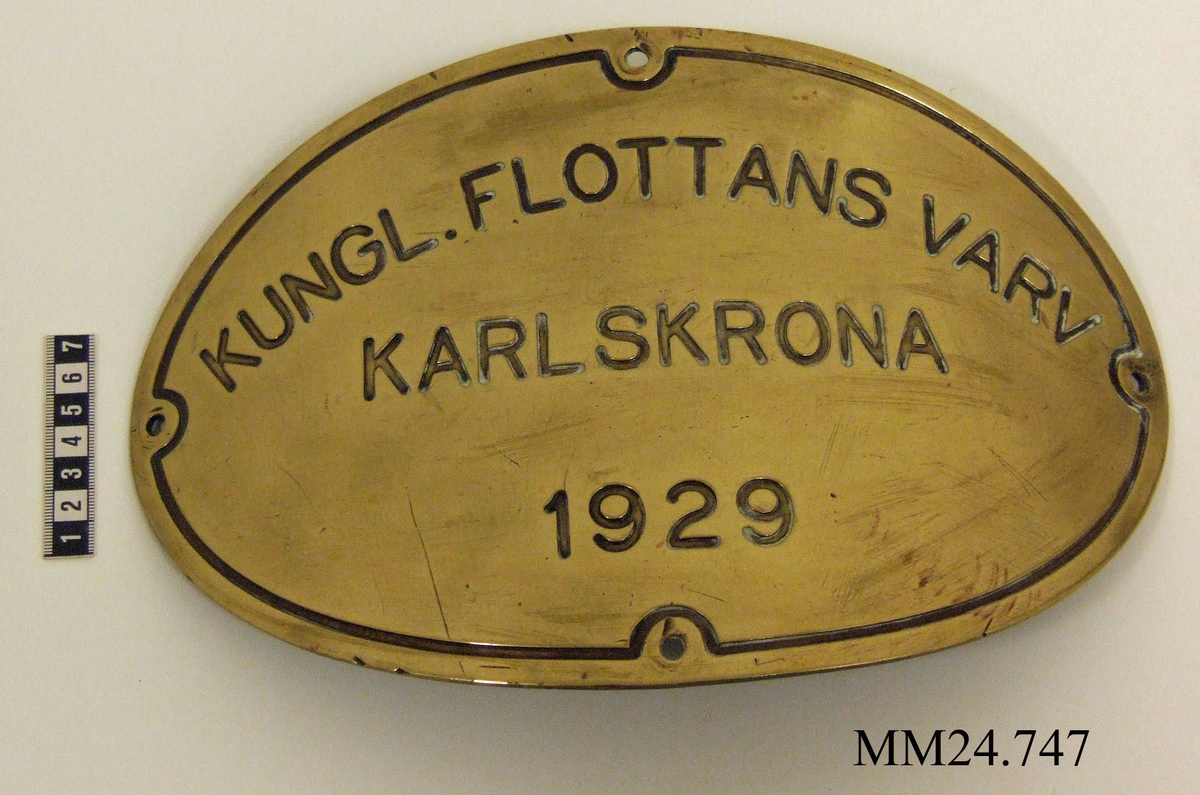 Oval metallskylt. Skylten  är böjd som för att fästas på någonting runt. Skruvhål mitt på varje lång- och kortsida. Text på skylten: "KUNGL.FLOTTANS VARV KARLSKRONA 1929".