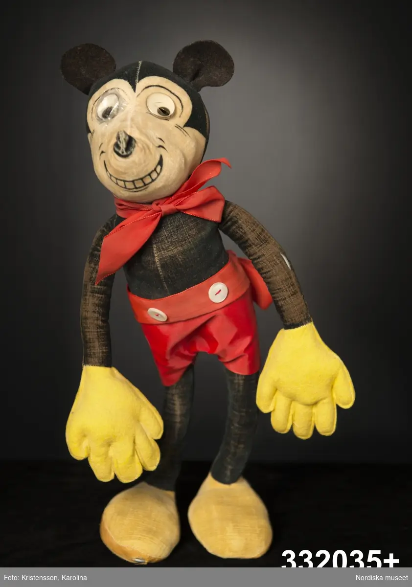 Docka av tyg, föreställande seriefiguren Musse Pigg, en mus klädd i röda kortbyxor och gula handskar och skor.  Lång och smal kropp med böjbara stoppade armar och ben. Stomme av ståltråd, klädd med mjuk stoppning och plysch. Svart kropp och runda musöron av filt, ansikte ljust beige med påklistrade ögon av plast, varav det ena är lagat med tejp och det andra har en spricka. Svart nos, karaktäristiskt leende med synliga tänder målade direkt på tyget. Gula skor och handskar, de senare av filttyg. Tyget är delvis bortnött så att den stålvajer som går genom dockans armar och ben är synlig. Dockan ser ut att vara lagad på några ställen. Det röda bandet med rosett om halsen kan vara ett senare komplement, liksom byxorna med två påsydda vita knappar i linningen.
/Karin Dern 2014-01-27