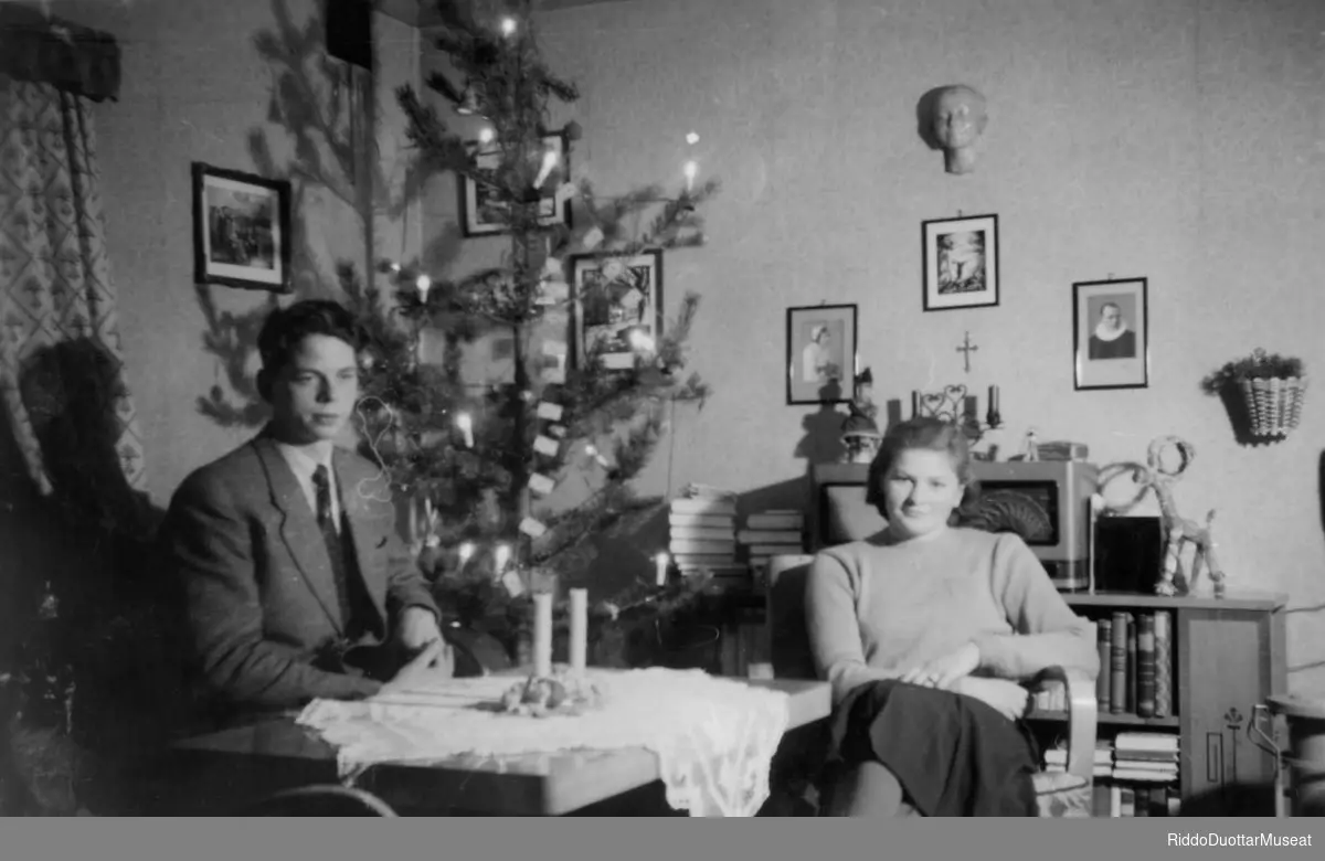 Bardni ja nieida cohkaba stobus, juovlamuorra duogábealde.
En mann og en kvinne i stuen, juletre i bakgrunnen.