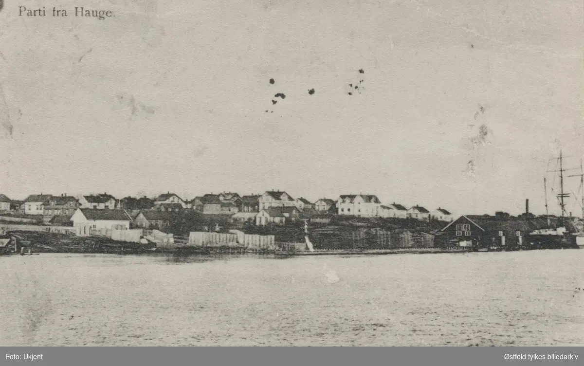 Bebyggelsen på Haugei Rolvsøy fotografert fra Glomma. Tl høyre ses et lagerhus og deler av to seileskuter. Pstkort.