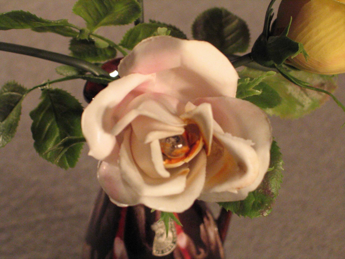 Lampe der foten er forma som ein vase med roser der det er sett små pærer i kvar av blomane. Slank konisk vase med hank og tut, oppi denne 2 raude, 2 rosa og 1 kvit plastrose.
Kvit plastledning med bryar midt på, truleg sekundær
