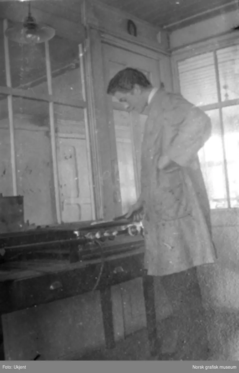 En ung mann i arbeidsfrakk står ved et arbeidsbord, sannsynligvis et elektrolysekar. Innendørs på Dreyers Reproduktionsanstalt i Stavanger. Tekst under bildet i albumet: "Elektrisk indbrænding".