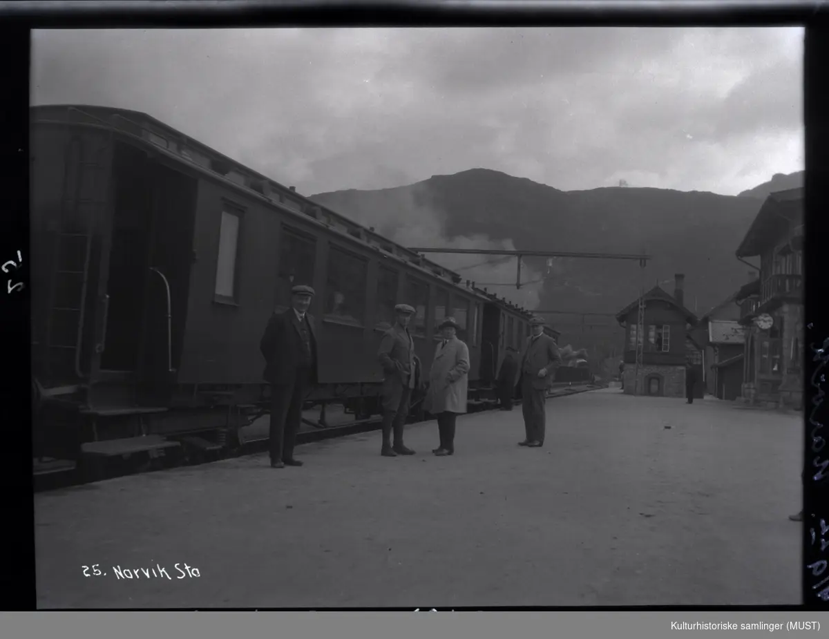 Fire menn på perrongen. 
Jernbanevogn og stasjonsbygningen på Narvik stasjon.