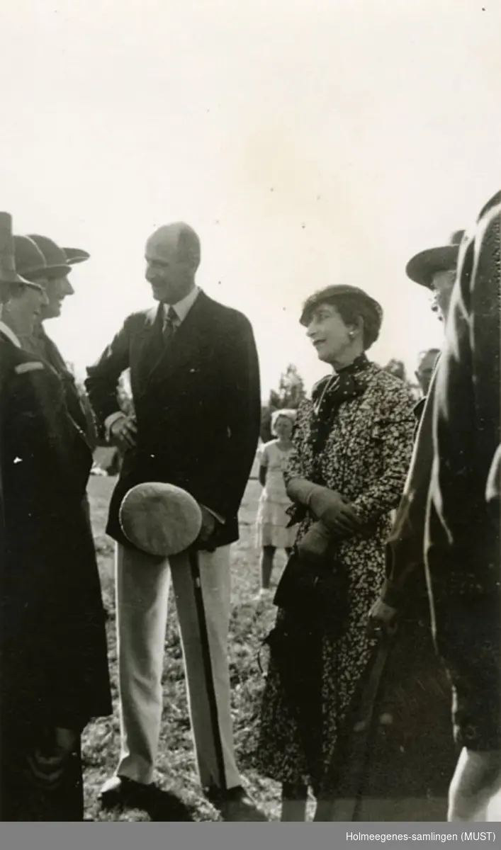 Kong Haakon VII og dronning Maud i samtale med noen kvinner på speiderleir i 1935. Håndskrevet på baksiden av fotografiet: "Kongen og dronningen snakker med engelske speiderpiker".