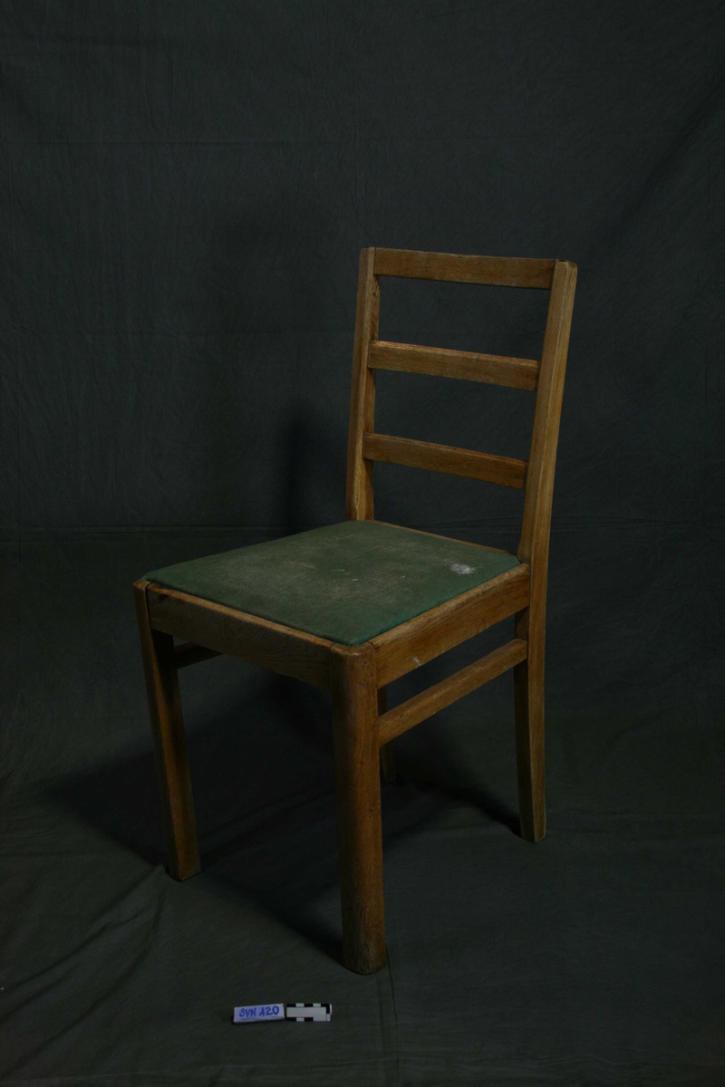 Stol med fire ben, og rygg med tre tverrgående treplanker. Setet er laget i grønt stoff, og har flekker. 
