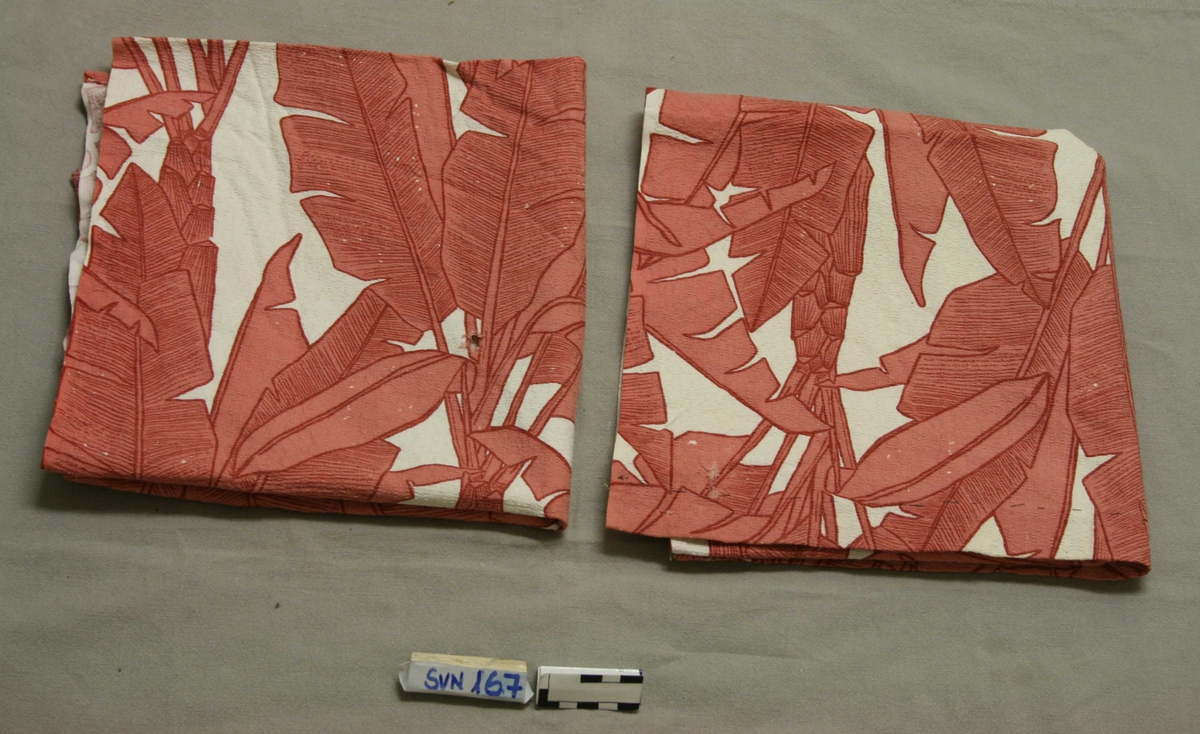 Rektangulær tekstilgardin med rød-rosa plantemønster (palmeblader) på hvit bunn. 