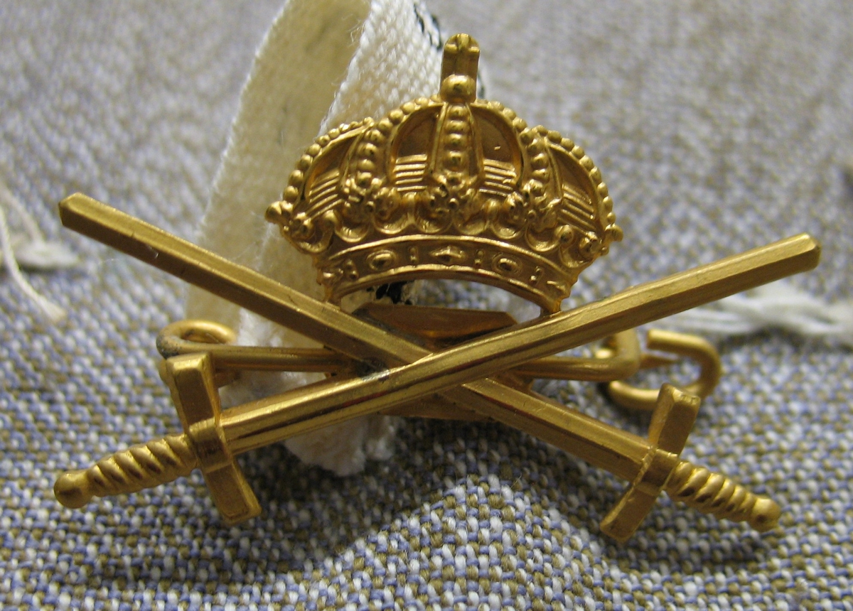 Arméns tecken i gulmetall med två korslagda svärd under en krona. Emblemet/märket sitter på sin originalplats på dräkten (21 553:1).

Övriga upplysningar se nr 21 553, samlingsblankett.