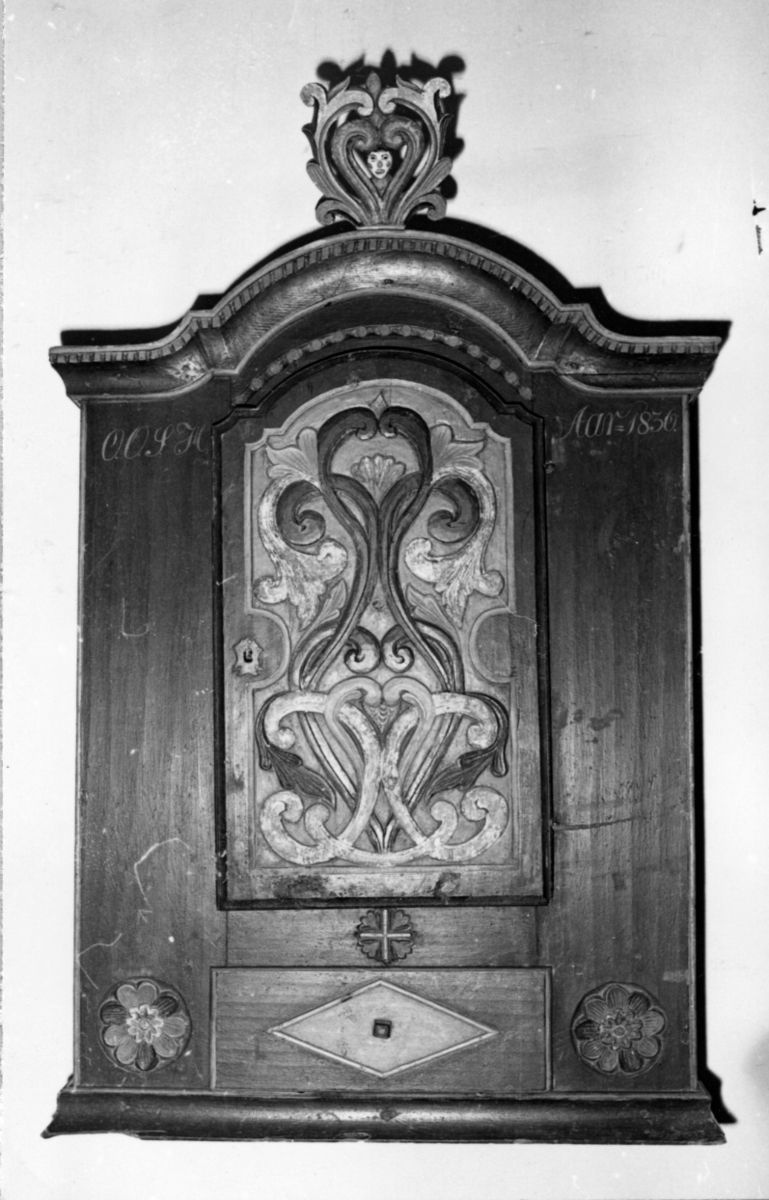 Skapet har en dør som er buet oventil og dekorert med utskåret dobbelranke i relief. Nederst har skapet en skuffe, på hver side av denne en utskåret rosett. Over skuffen en mindre rosett