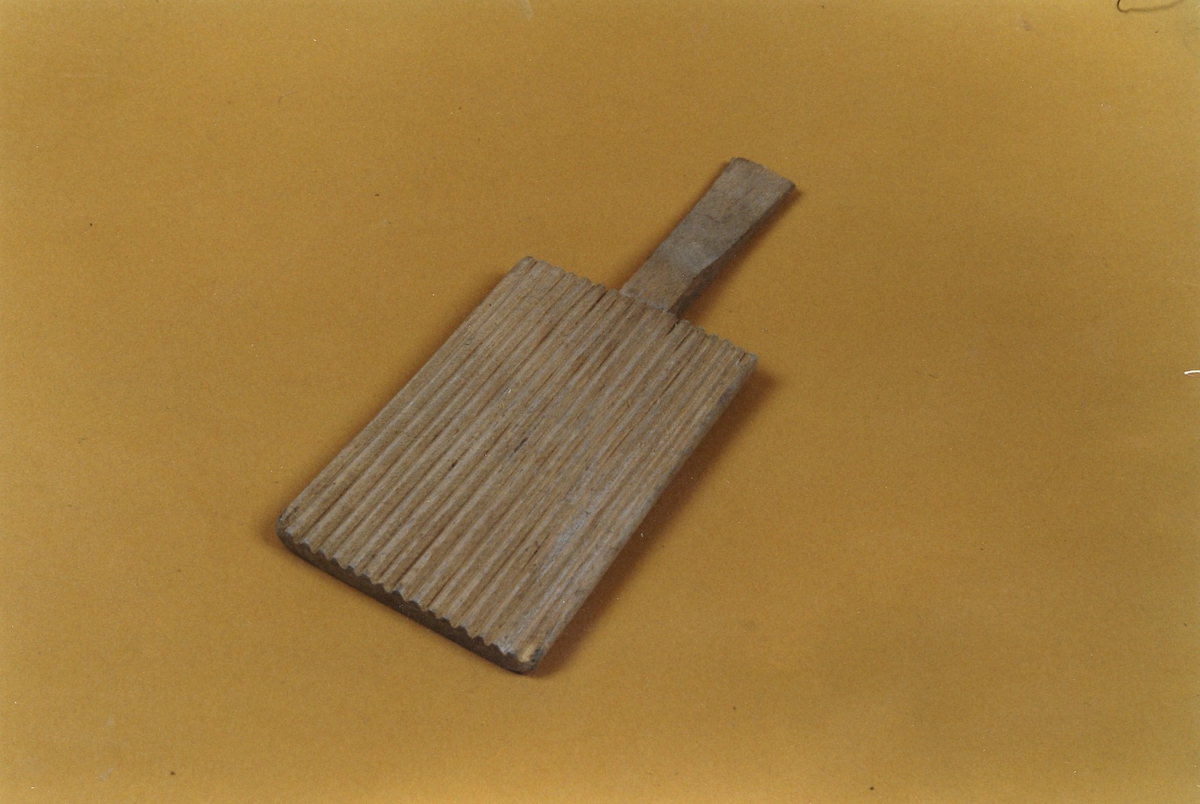 Smørspade i ubehandlet tre, hvor spadebladet er rektangulært med tynne riller og håndtaket er smalt og flatt. 